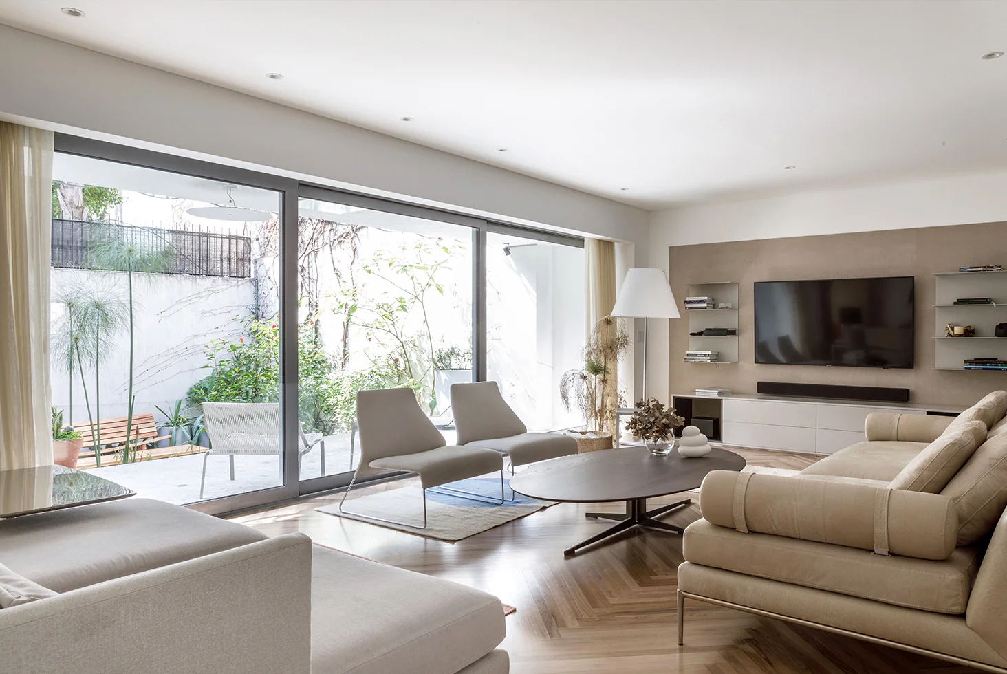 Modern living room with large sliding glass doors opening to a garden, featuring beige sofas, contemporary chairs, and a wall-mounted TV.